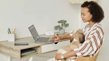 Kadın evde laptopta çalışırken köpeğini kucaklıyor.